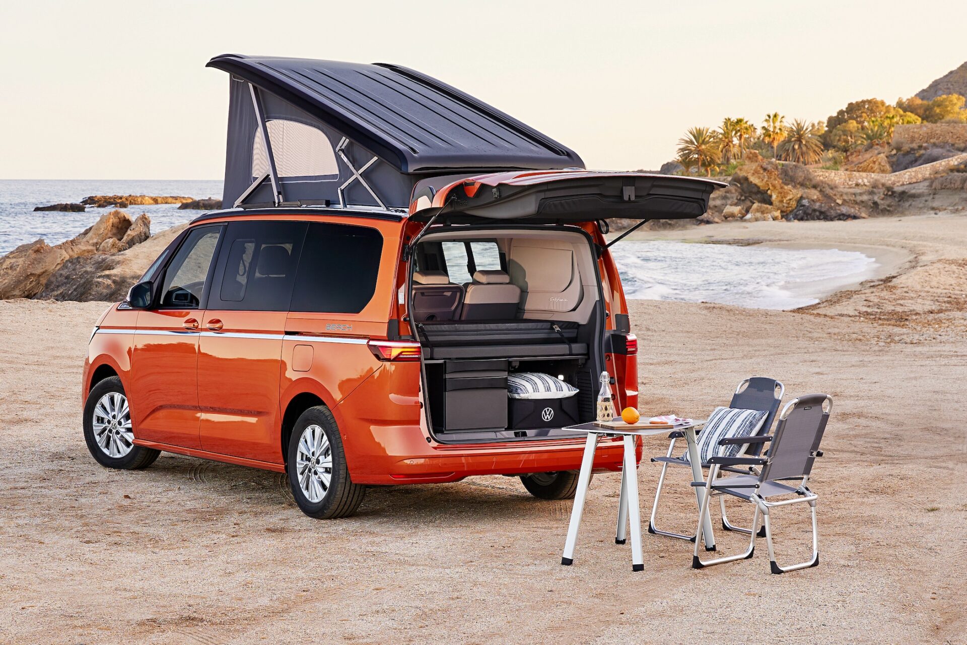 Orange Volkswagen husbil parkerad på stranden med öppen baklucka som visar campingutrustning och ett bärbar kök.