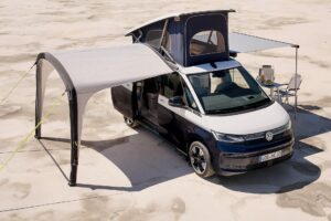 Volkswagen husbil med utsträckt sidomarkis över en uppsättning utomhusstolar och bord, parkerad på en texturerad ökenyta.