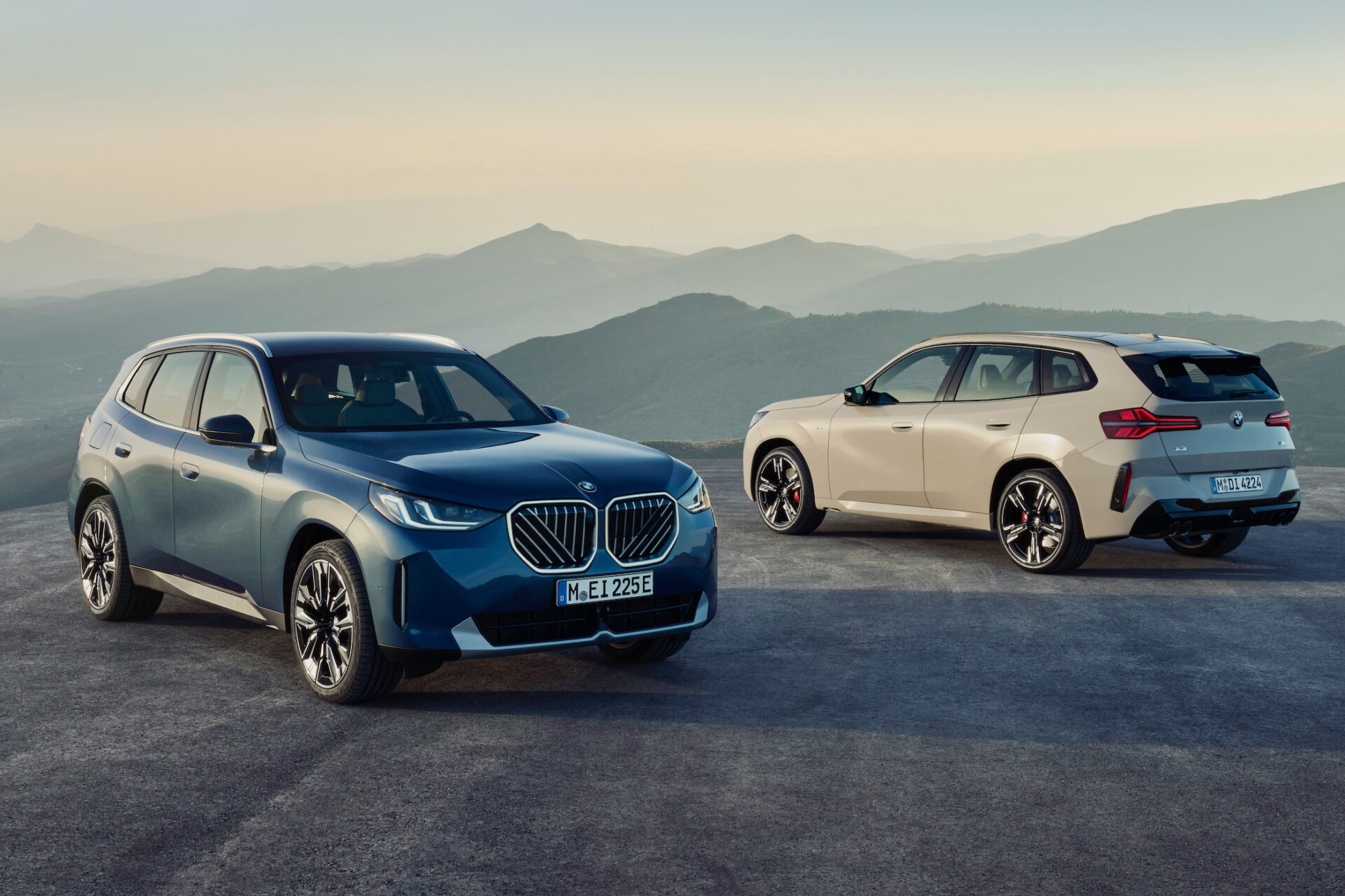 Två BMW SUV , en blå och en beige, står parkerade på en väg med berg i bakgrunden.