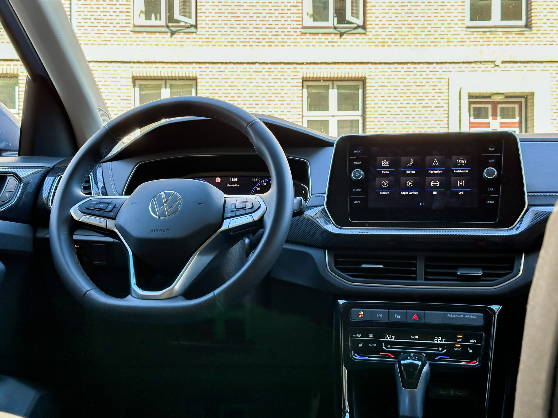 Interiör av Volkswagen med ratt och pekskärm, byggnad synlig genom fönstret.
