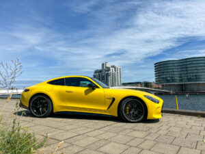 Gul Mercedes-AMG GT parkerad på en kaj med moderna byggnader i bakgrunden.