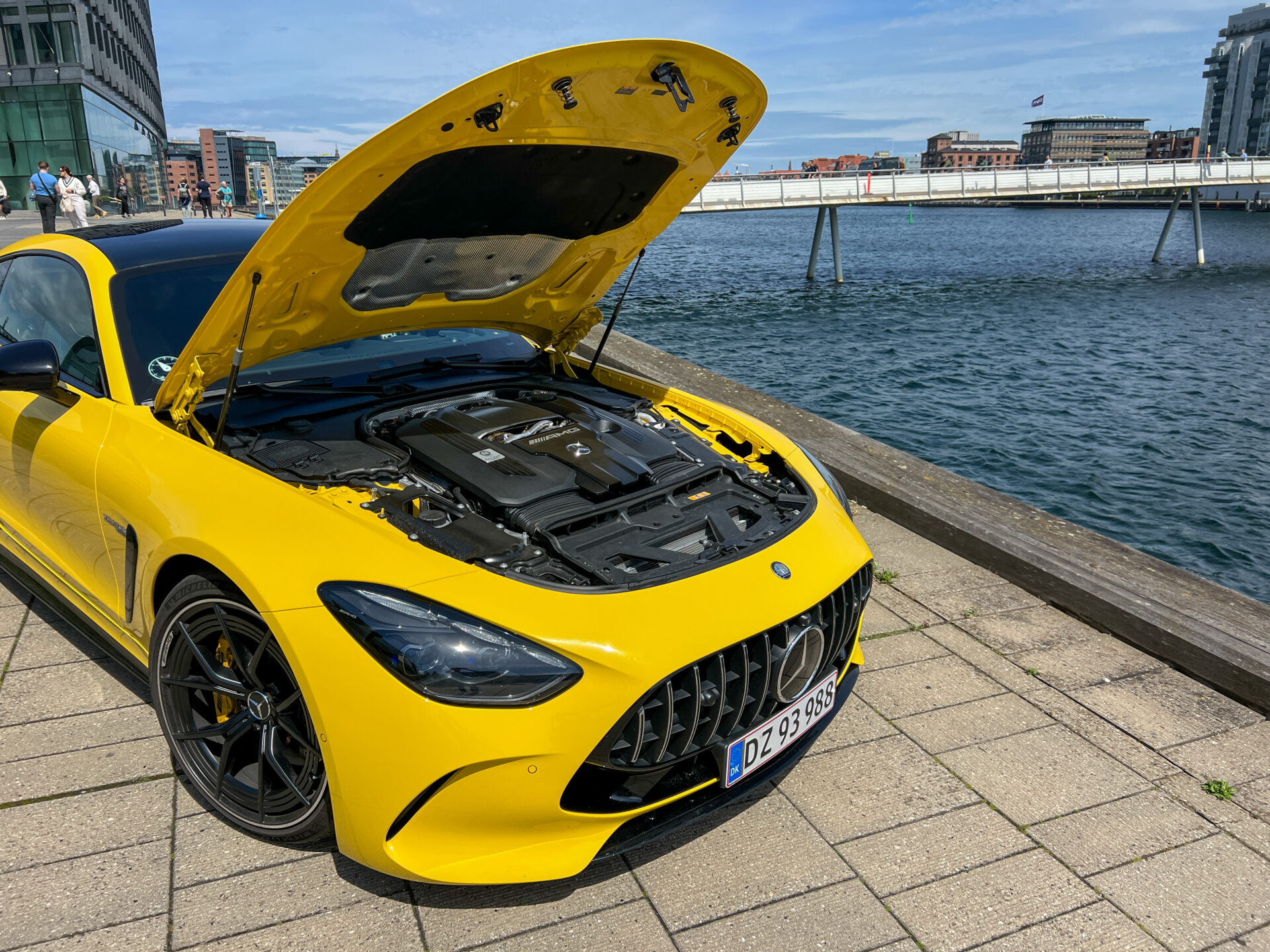 Gul Mercedes-AMG GT med öppen motorhuv på en kaj vid vattnet.