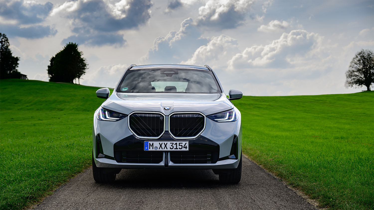 Frontal vy av en silverfärgad BMW SUV som står på en landsväg, omgiven av gröna fält och träd i bakgrunden.