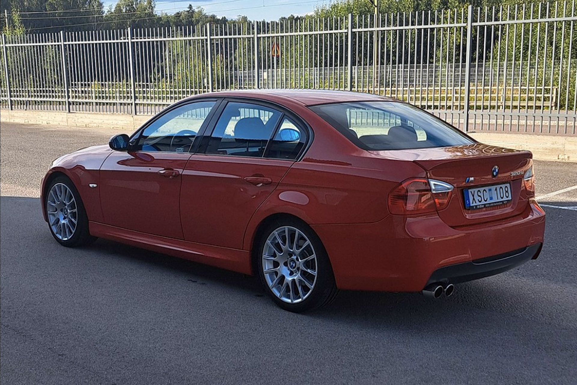 Röd BMW sedan sedd bakifrån, parkerad vid ett staket.