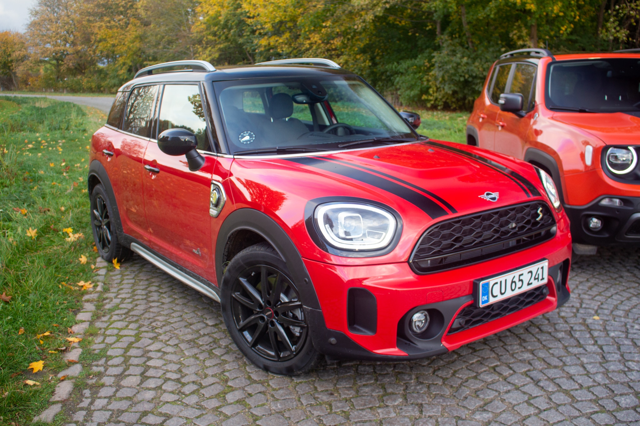 Rödsvart mini countryman parkerad på uppfart bredvid orange bil.